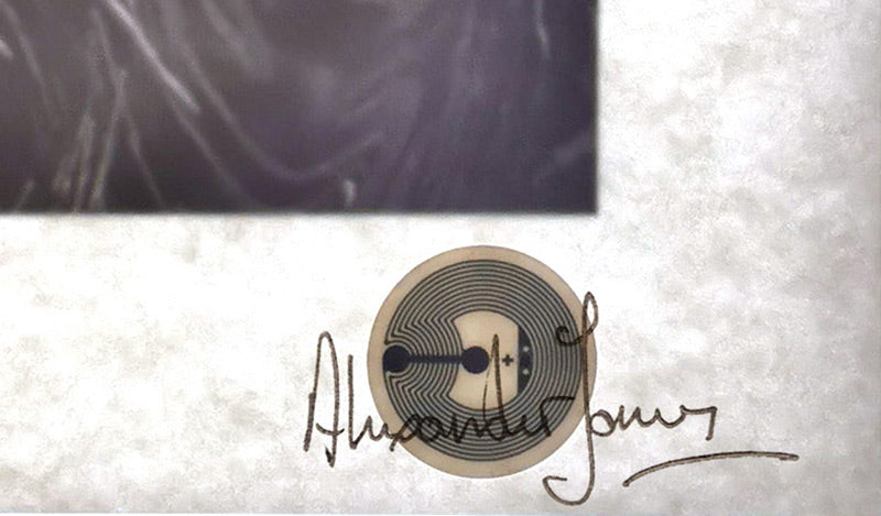 A round NFC seal under the paper of a print, behind the artist's signature, seen against the light